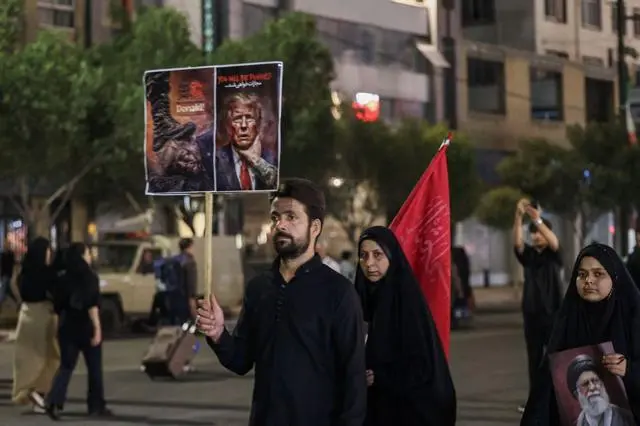 شاب يرتدي قميصاً أسود وشعره أسود كثيف، ولديه لحية سوداء خفيفة تغطي وجهه، بينما يحمل لافتة مناهضة للولايات المتحدة لدى وصوله مع آخرين لحضور مراسم تشييع الزعيم الإيراني الراحل علي خامنئي وأفراد من عائلته بالقرب من مرقد الإمام الرضا في مدينة مشهد، في وقت مبكر من يوم 9 يوليو/تموز عام 2026. ويستعد الإيرانيون اليوم لدفن زعيمهم الراحل علي خامنئي في مسقط رأسه بمدينة مشهد، شمال شرق إيران وتأتي مراسم دفن المرشد الأعلى الراحل عقب جنازة استمرت عدة أيام وجذبت ملايين المشيعين في مدن مختلفة بإيران والعراق.