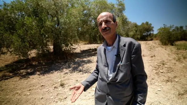 أبو مضيف تظهر صورته من الأعلى مرتدياً سترة بدلة رسمية رمادية اللون، في جيبها قلم حبر أزرق، ويرتدي قميصاً مخططاً بالأبيض والأزرق.. كبير في السن وبشرته سمراء وفقد نصف شعر رأسه ولديه شارب دون لحية. ولون شعره أسود فيه خصل بيضاء. يحرك يده متحدثاً وهو يقف على أرض خلفها أشجار. الصورة التقطت نهاراً.