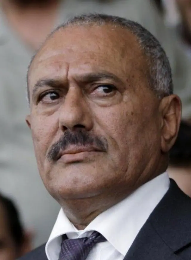 علي عبدالله صالح في صورة تظهر وجهه ذو البشرة السمراء وشاربه وشعره الذي يغزوه الشيب، مرتدياً سترة بدلة رمادية اللون مع ربطة عنق بنفس اللون لكن منقطة بلون رمادي أفتح مع قميص أبيض.