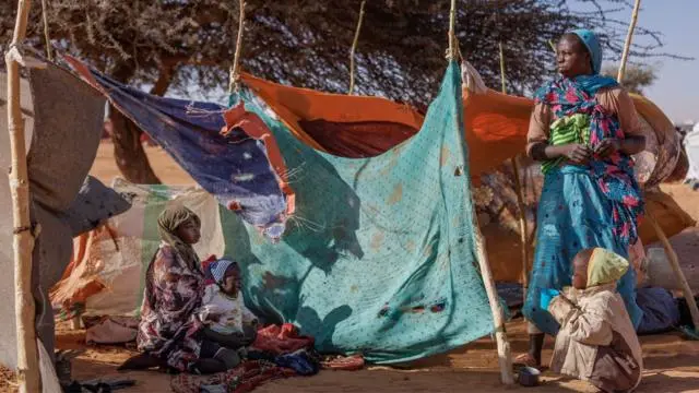 امرأتان وطفلان — ينحدرون أصلاً من مدينة الفاشر — يقفون قرب قطع قماش مربوطة بعصيّ تُستخدم كمأوى مؤقت في تشاد
