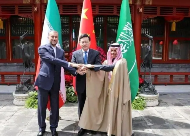 وزير الخارجية الإيراني حسين أمير عبد اللهيان ووزير الخارجية السعودي الأمير فيصل بن فرحان آل سعود ويتوسطهما وزير الخارجية الصيني تشين غانغ يضعون أيديهم فوق أيدي بعضهم البعض مصافحين، خلال اجتماع في بكين. وخلفهم أعلام بلادهم.
