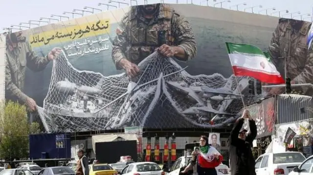 لافتة كبيرة معلقة في شارع في طهران كتب عليها: "سيبقى مضيق هرمز مغلقاً؛ والخليج بأكمله هو ساحة صيد لنا".
رجل يحمل العلم الإيراني وسيدة ترتدي وشاح العلم الإيراني وتحمل صورة المرشد مجتبة الخامنئي