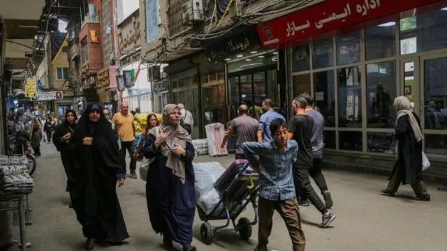 طفل إيراني يجر عربة بضائع صغيرة في أحد الأسواق، ويمر من جانبه سيدتان محجبتان وخلفه عدد من الأشخاص.