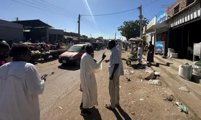 صورة لشارع في مدينة الأبيض به سيارة وباعة جائلون، وسيارة. بينما يقف رجل يرتدي جلباباً أبيض يتحدث إلى رجل يرتدي قميصاً وسرولاً أبيض لونهما رمادي.