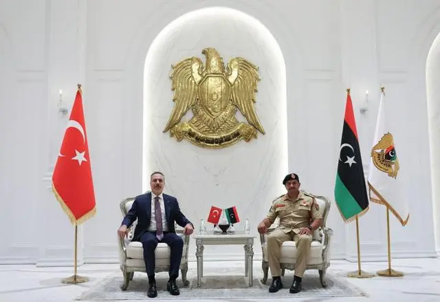 صدام حفتر يرتدي لباساً عسكرياً ويجلس على كرسي وفي المنتصف علما تركيا وليبيا صغيرا الحجم على طاولة صغيرة وفي الجانب الأيسر هاكان فيدان يرتدي بدلة رسمية كحلية اللون مع قميص أبيض وربطة عنق تبدو كحلية اللون منقطة بالبنفسجي، وخلفهما العلم الليبي والعلم التركي في قاعة يغلب عليها اللون الأبيض.